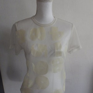 Peut inclure: T-shirt en maille filet blanche avec un imprimé graphique crème du mot "OUT OF ORDER" en lettres rondes et de grande taille.