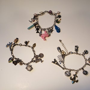 Könnte beinhalten: Drei silberne Armbänder mit verschiedenen Charms, darunter ein rosa Flamingo, ein Eiffelturm, eine Palme, eine Weinflasche, eine Fleur-de-lis und ein Zebra-Charm.