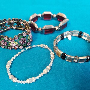 Pode incluir: Quatro pulseiras de vários estilos são exibidas em uma superfície azul. Uma apresenta pedras vermelhas retangulares, outra tem desenhos florais com detalhes roxos e verdes, uma terceira tem cristais transparentes e a última tem pedras retangulares pretas e transparentes.