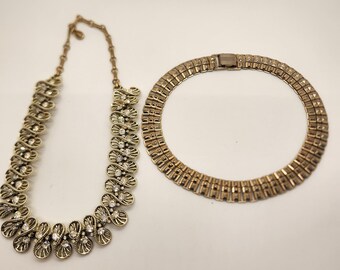 May include: Two gold necklaces, one with a geometric design and the other with a more ornate design featuring clear rhinestones.