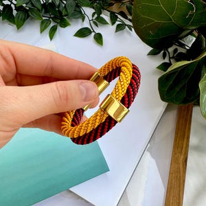 May include: A black and red braided bracelet with a gold magnetic clasp. The bracelet is made of two strands of braided cord, one black and one red, that are twisted together. The gold clasp is a simple, rectangular design.