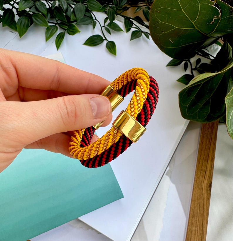 May include: A black and red braided bracelet with a gold magnetic clasp. The bracelet is made of two strands of braided cord, one black and one red, that are twisted together. The gold clasp is a simple, rectangular design.