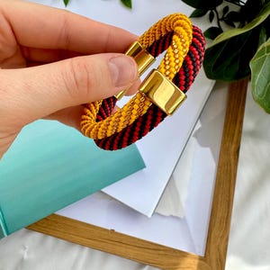 May include: A black and red braided bracelet with a gold magnetic clasp. The bracelet is made of two strands of braided cord, one black and one red, and the strands are intertwined.
