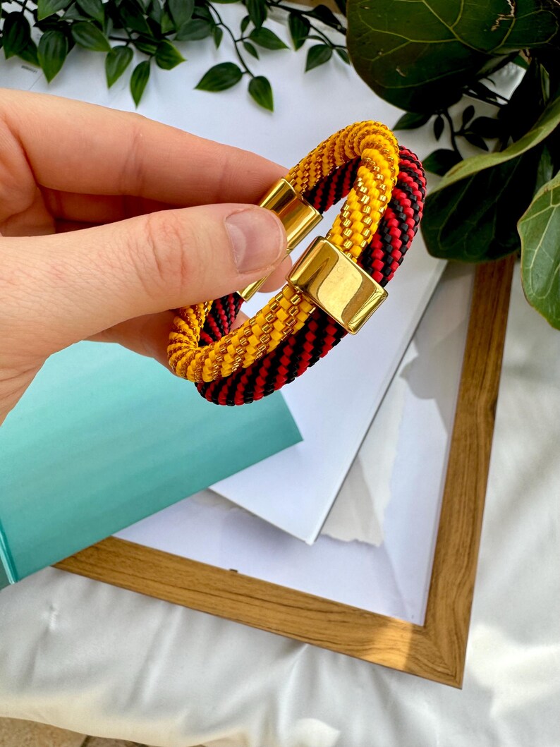 May include: A black and red braided bracelet with a gold magnetic clasp. The bracelet is made of two strands of braided cord, one black and one red, and the strands are intertwined.