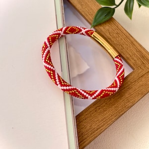 May include: Red, white, and gold beaded bracelet with a geometric pattern. The bracelet has a magnetic clasp.