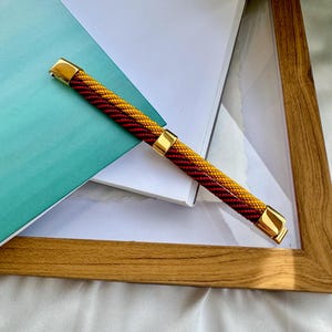 May include: A braided watch band with gold accents. The band is red and yellow.