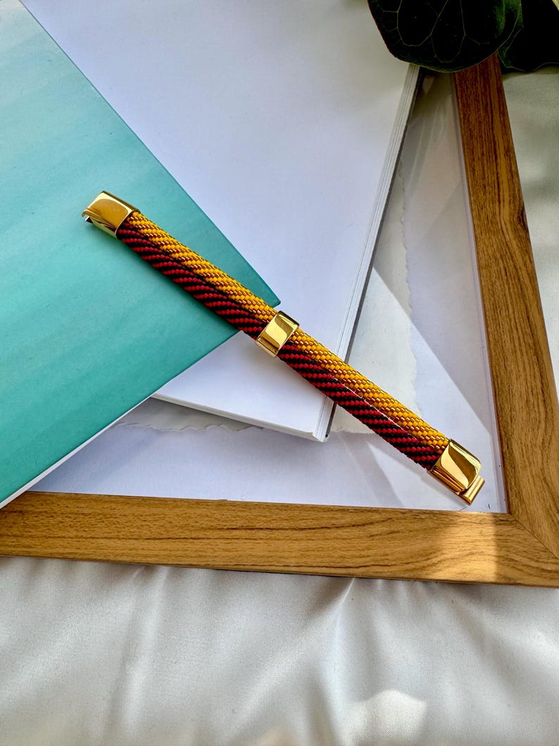 May include: A braided watch band with gold accents. The band is red and yellow.