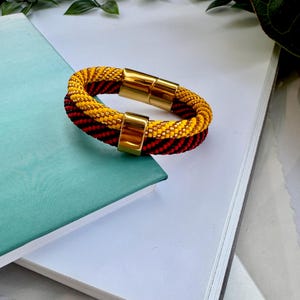 May include: A yellow and black beaded bracelet with a gold magnetic clasp. The bracelet is made of small beads and has a woven design.