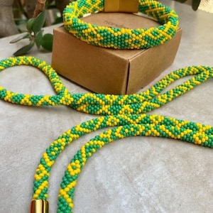 Op de afbeelding: Een groene en gele kralen ketting en armband set. De ketting is lang en heeft een gouden sluiting. De armband is korter en heeft een magnetische sluiting.