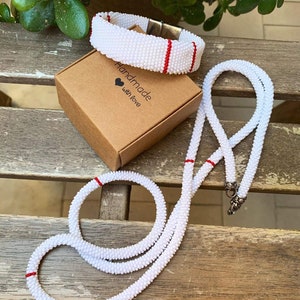 Può includere: Set di collana e braccialetto in perle bianche con dettagli rossi. Il set è esposto su una superficie in legno con una piccola scatola marrone che dice "Fatto a mano con amore".