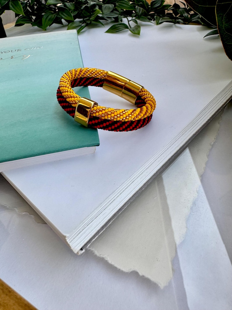 May include: A yellow and red beaded bracelet with a gold magnetic clasp. The bracelet is lying on a white surface.
