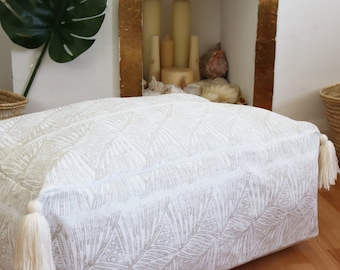 Op de afbeelding: Een wit en beige vloerkleed met kwastjes. Het kussen ligt op een houten vloer voor een met goud geverfde open haard.