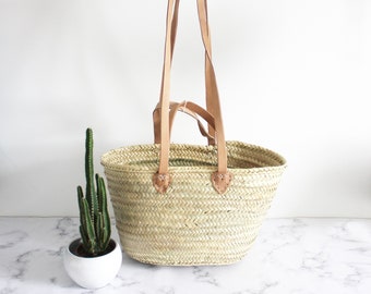 Peut inclure: Un sac cabas en paille tressée naturelle avec des anses et des sangles en cuir marron. Le sac est de forme ovale et a une large ouverture. Le sac est posé sur une surface en marbre blanc.