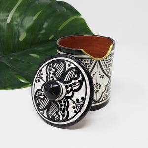 Könnte beinhalten: Eine schwarze und weiße Keramikdose mit Deckel. Die Dose hat ein geometrisches Muster und der Deckel ein Blumenmuster. Die Dose hat einen goldenen Rand.