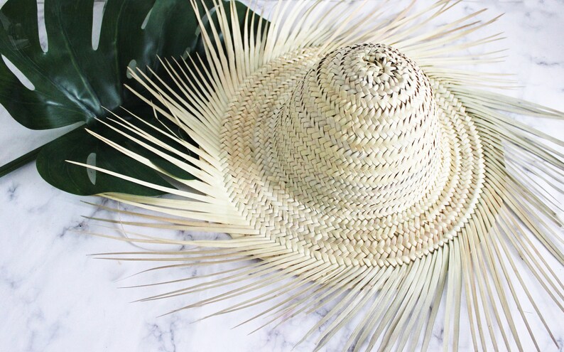 May include: A woven straw hat with a wide brim and a rounded crown. The hat is light beige in color and has a natural, textured appearance. A large, dark green monstera leaf is visible in the background.