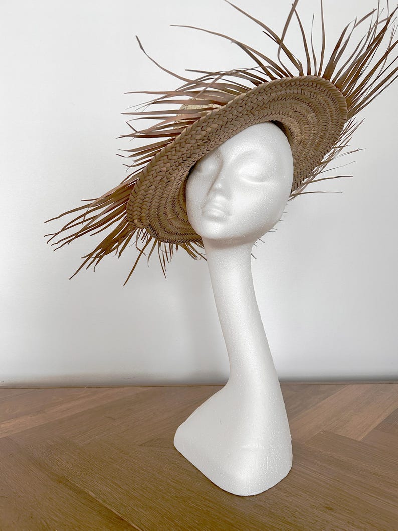 May include: A straw hat with a wide brim and decorative dried palm leaves. The hat is displayed on a white mannequin head and neck. The hat is a light brown color and has a woven texture.