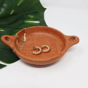 Puede incluir: Un pequeño plato de terracota con un acento dorado y dos pendientes de aro dorados en su interior. El plato tiene dos asas a los lados.