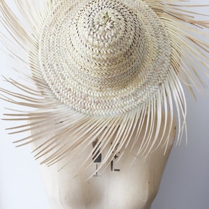 May include: A woven straw hat with a wide brim and a sunburst design. The hat is light beige and has long, thin straw extensions radiating from the brim. The hat is displayed on a mannequin.
