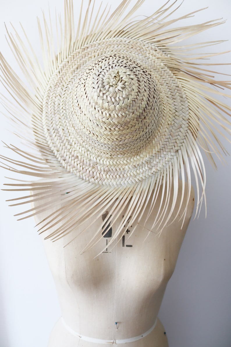 May include: A woven straw hat with a wide brim and a sunburst design. The hat is light beige and has long, thin straw extensions radiating from the brim. The hat is displayed on a mannequin.