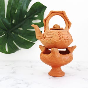 May include: A terracotta teapot and stand, with a handle and patterned detailing. The teapot rests on a pedestal base, with a large green leaf behind it. The pottery is a warm, earthy colour, typical of terracotta.