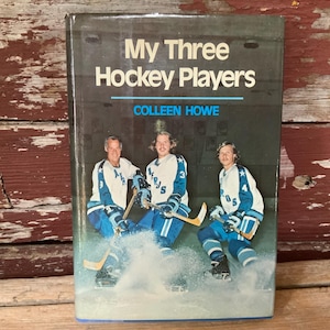 May include: A vintage book cover titled "My Three Hockey Players" by Colleen Howe. The cover features three hockey players in blue and white uniforms, skating on ice. The background is a dark, textured surface.