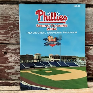 May include: A blue and white program for the Philadelphia Phillies Spring Training 2004. The cover features the team logo and a photo of the Bright House Field baseball stadium. The program is priced at $3.00.
