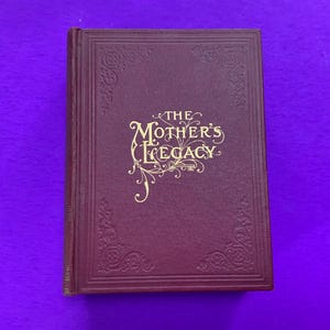 May include: A burgundy-colored book titled "The Mother's Legacy" with gold lettering and decorative corner designs. The book has a textured cover and is set against a purple background.