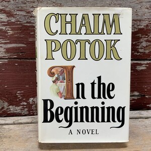 May include: A hardcover book titled "In the Beginning" by Chaim Potok. The book cover is white with black and gold lettering. An illustration of two men at a table is featured on the cover. The book is a novel.