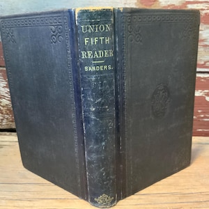 May include: An antique book titled "UNION FIFTH READER" by Sanders. The book has a dark blue cover with embossed decorative patterns and text in gold lettering. The spine is a lighter blue with the title and author's name. The book is displayed open.