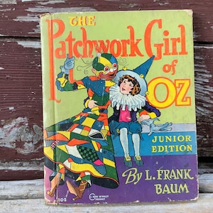 May include: A vintage book cover for "The Patchwork Girl of Oz" by L. Frank Baum. The cover features a colorful illustration of a patchwork girl and a jester-like figure. The book is a junior edition.