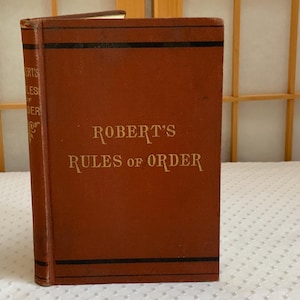 May include: A hardcover book titled Robert's Rules of Order with a brown cover and black horizontal stripes. The title is printed in gold lettering on the front cover and the spine. The book is standing upright with visible wear on the edges.