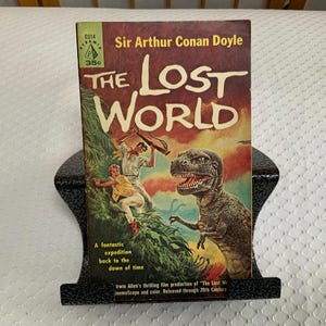May include: Vintage paperback book titled "The Lost World" by Sir Arthur Conan Doyle. The cover features a colorful illustration of a dinosaur and people in a jungle setting. The book is displayed on a black metal book stand.