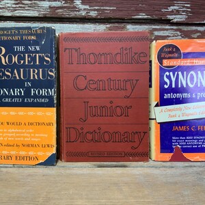 May include: Three vintage hardcover books on a wooden surface. The book on the left is blue with gold lettering and reads "The New Roget's Thesaurus in Dictionary Form". The middle book is red with black lettering and reads "Thorndike Century Junior Dictionary". The book on the right is blue with orange and gold lettering and reads "Funk & Wagnalls Standard Handbook of Synonyms, antonyms & prepositions".
