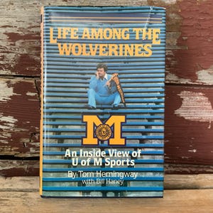 May include: A book cover with a blue and yellow striped background. The title is "Life Among the Wolverines: An Inside View of U of M Sports" by Tom Hemingway with Bill Haney. A man in a blue sweater is sitting on the bleachers.