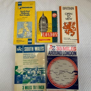 May include: A collection of five vintage paper road maps and travel guides featuring titles such as Michelin South East Midlands East Anglia, London, Britain Open to View, RAC South Wales, and A-Z 35 Miles Around London with various illustrations.