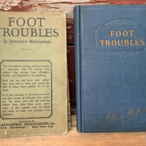 May include: Two vintage books titled "Foot Troubles" by Bernarr Macfadden. The book on the left has a green cover with black text. The book on the right has a blue cover with gold text. The book on the right has a signature on the bottom right corner.