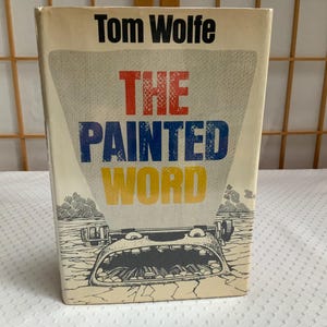 May include: A hardcover book titled The Painted Word by Tom Wolfe. The cream-colored dust jacket features the author's name in black at the top, with the title in red, blue, and yellow block letters above a black and white illustration of a typewriter.