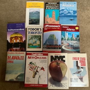 May include: A collection of travel guide books with various destinations, including Alaska, Mexico City, Acapulco, San Francisco, Toronto, Montreal, Quebec City, Atlanta, Hawaii, New Orleans, New York City, and Texas. The books are arranged in two rows on a brown carpet.