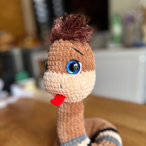 May include: A brown crocheted snake toy with a red tongue sticking out. The snake has blue eyes and brown hair on its head.