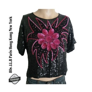 Könnte beinhalten: Ein schwarzes Paillettenoberteil mit einem rosafarbenen Blumendesign. Das Oberteil ist kurz und hat kurze Ärmel. Der Text "80s J.L.B Paris Hong Kong New York" steht auf der linken Seite des Bildes.