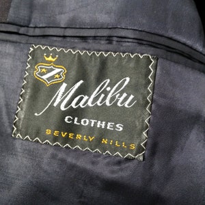 May include: A black label with white stitching and the text "Malibu Clothes Beverly Hills" in white lettering. A gold crown and shield with stars are above the text.