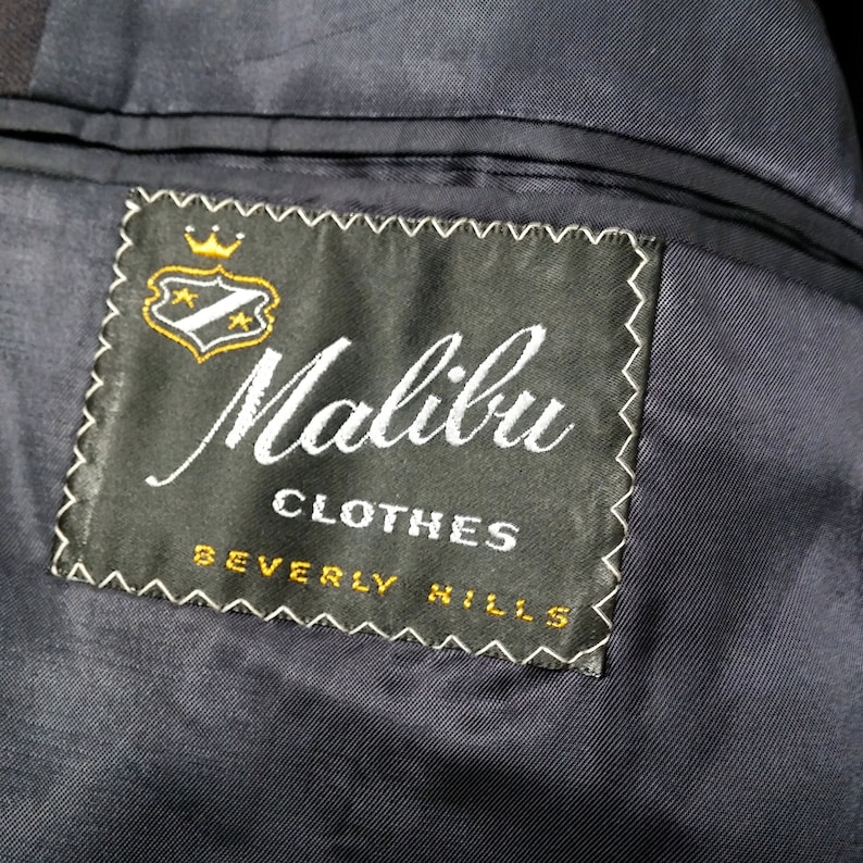 May include: A black label with white stitching and the text "Malibu Clothes Beverly Hills" in white lettering. A gold crown and shield with stars are above the text.