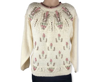 Può includere: Un maglione di lana color crema con un motivo floreale rosa e verde. Il maglione ha un collo rotondo e maniche lunghe.