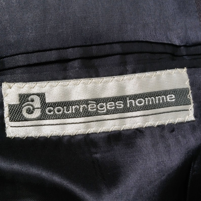 May include: A white label with black stitching and the text "Courr&egrave;ges homme" in black lettering.