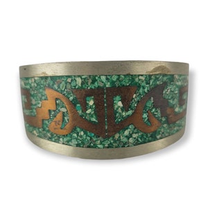 Op de afbeelding: Een zilveren armband met een groen en koperen inlegontwerp. Het ontwerp heeft een herhalend patroon van gestileerde vormen.