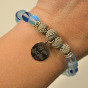 Op de afbeelding: Een armband met blauwe en witte glazen kralen, grijze lavasteen kralen en een zilveren bedel met de tekst "Laat je licht schijnen".