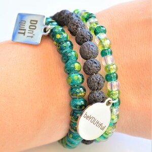 May include: Three bracelets stacked on a wrist. The top bracelet is black lava rock beads. The middle bracelet is green glass beads with gold flecks. The bottom bracelet is green and white iridescent beads. The bracelets have two silver charms, one that says "Don't quit" and the other that says "beYOUtiful".