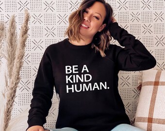 May include: A woman wearing a black sweatshirt with the text "BE A KIND HUMAN." in white lettering. She is sitting on a white couch with a brown and white plaid throw pillow behind her.