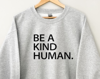 May include: A light gray sweatshirt with the text "BE A KIND HUMAN" in black. The sweatshirt is hanging on a wooden hanger against a white background. The sleeves are cuffed, revealing a white interior.