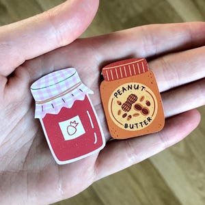 Puede incluir: Dos imanes de madera con forma de tarro. Uno es rojo con un diseño de fresa y una tapa de cuadros rosas. El otro es marrón con una etiqueta de mantequilla de cacahuete y un gráfico de cacahuetes. Los imanes se sostienen en una mano abierta.